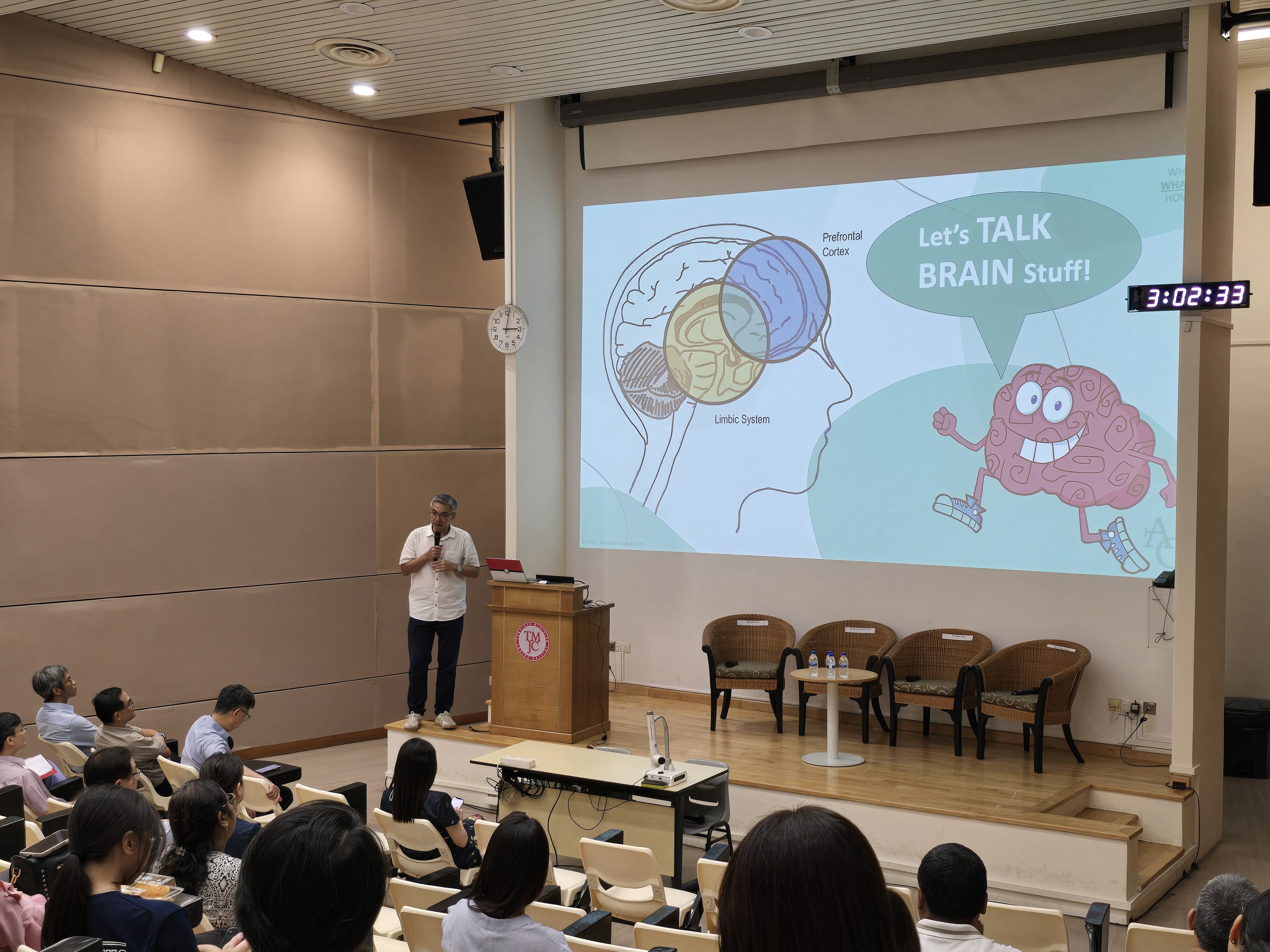 A male speaker stands at a podium in a lecture theatre, addressing an audience of parents seated in tiered rows. The large projected screen behind him shows a colourful slide featuring a diagram of the human brain with labels for the Prefrontal Cortex and Limbic System, alongside a cartoon brain character and the text "Let's TALK BRAIN Stuff!" A digital timer on the wall reads 3:02:33.